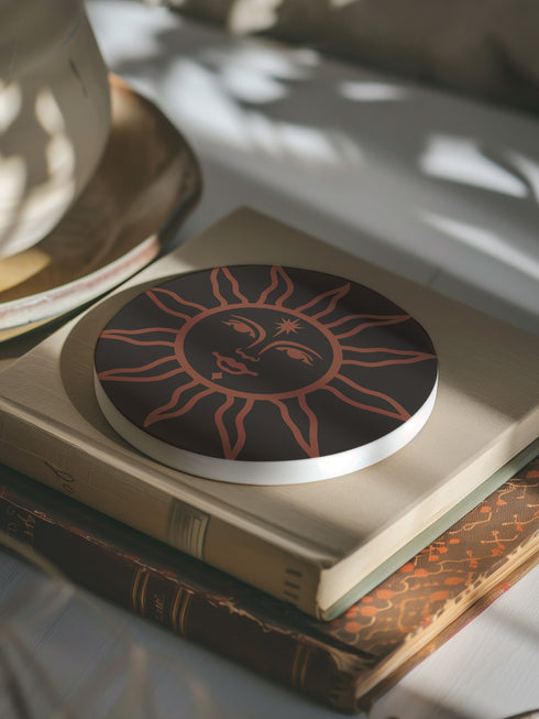 Round ceramic coaster in deep black featuring an intricate burnt-orange sun illustration with a serene, symmetrical face, radiating curved rays, and a delicate star detail on the forehead. The smooth, matte surface enhances the contrast between the dark background and the warm-toned design, creating a celestial, vintage-inspired aesthetic.